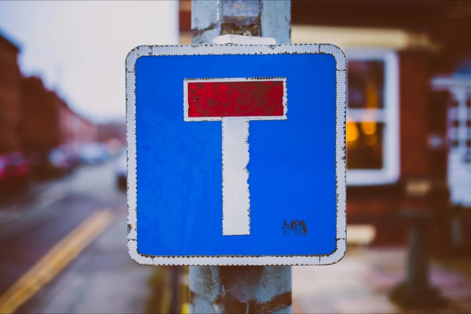 A blue no-through-road sign (red bar over a white T) on a pole in a terraced street